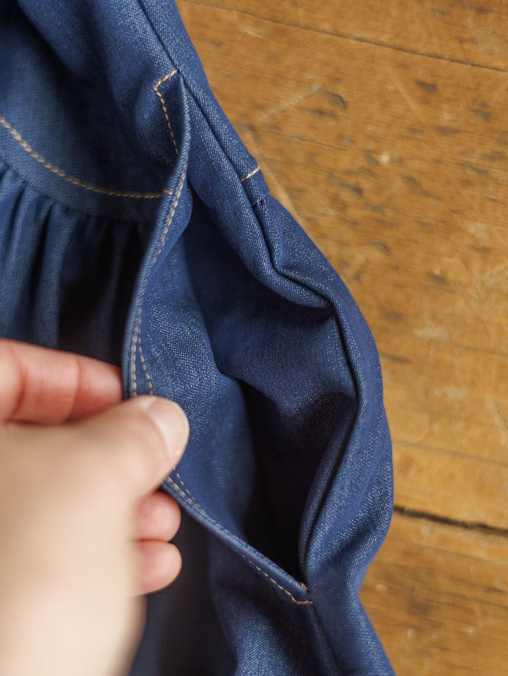 A hand opens the pocket of The Navassa, a blue denim midi dress with tan stitching by Savannah Sevens western life{&}style, against a wooden background.
