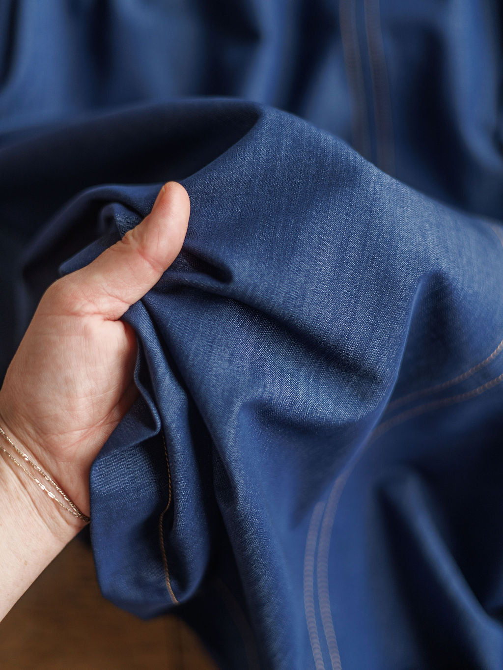 A hand gently grasps the soft blue denim fabric of The Navassa midi dress by Savannah Sevens western life{&}style, highlighting its texture and subtle stitching.