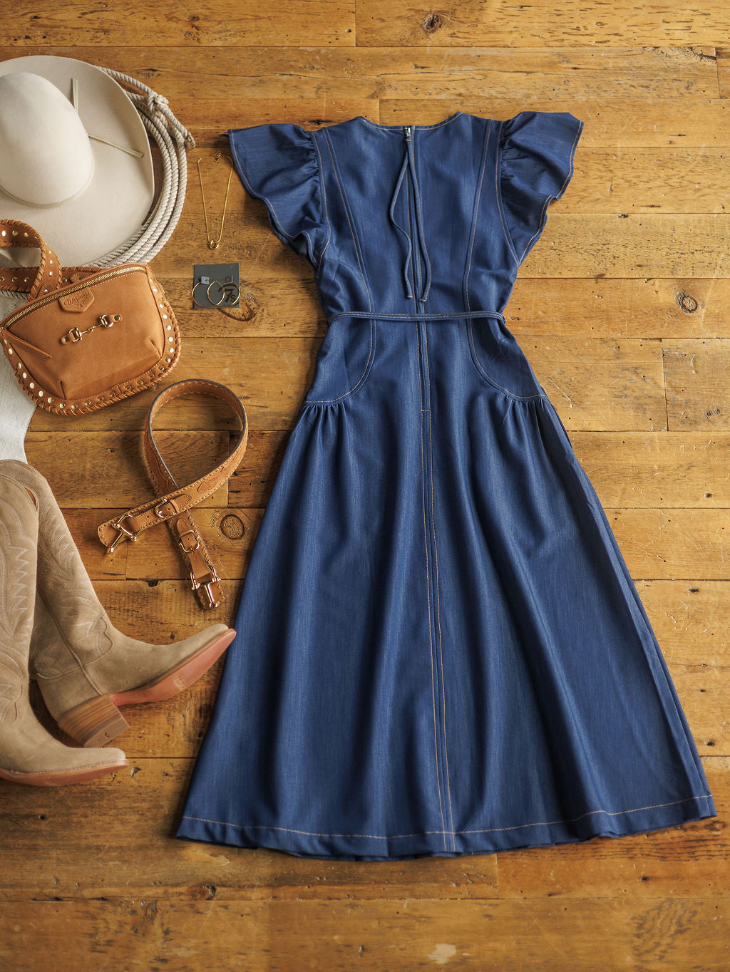 The Navassa denim midi dress by Savannah Sevens western life{&}style is styled on a wooden floor with tan cowboy boots, a brown studded bag, wide belt, silver necklace, and hat.