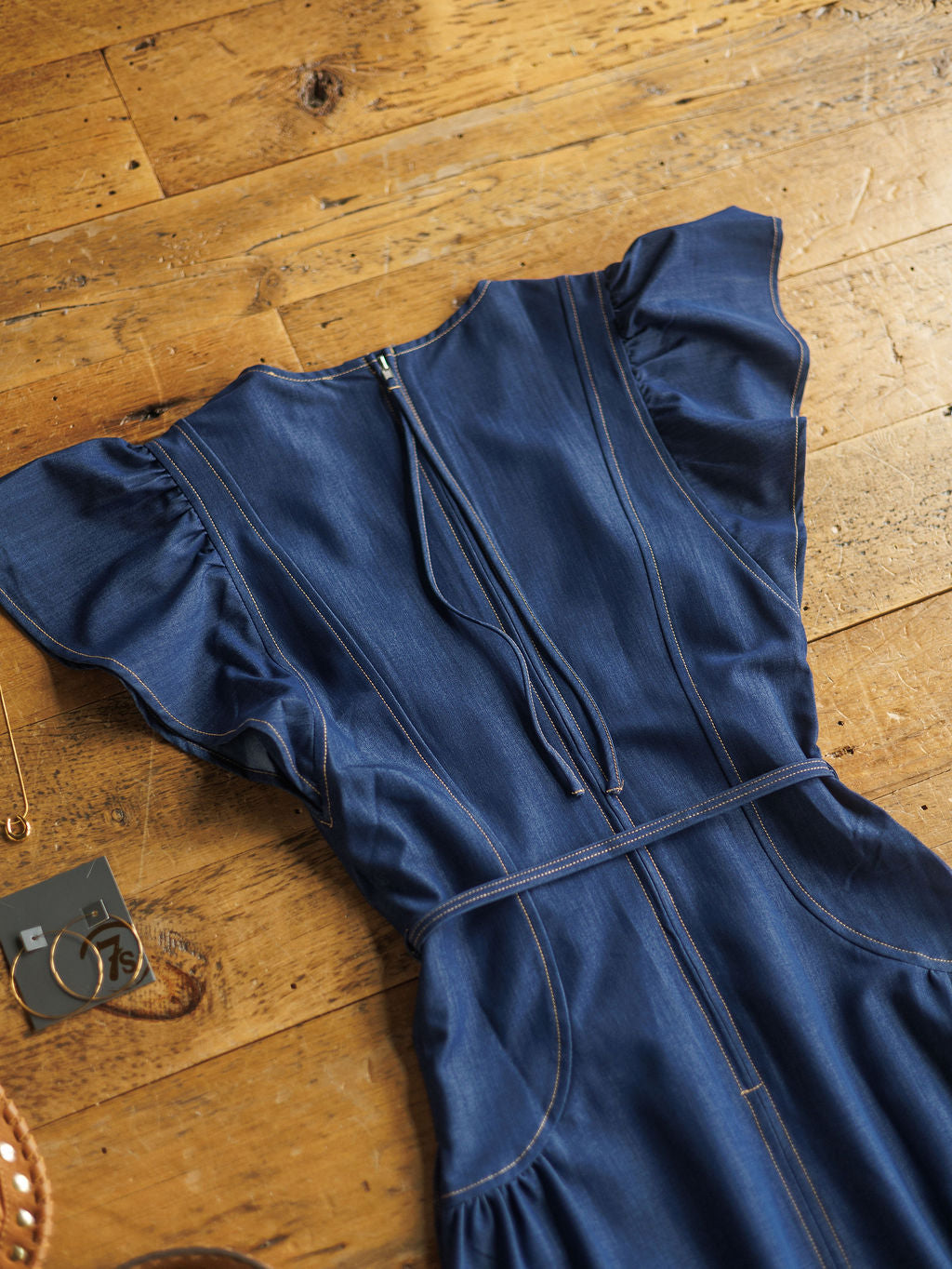 The Navassa blue denim midi dress from Savannah Sevens western life{&}style, featuring short flutter sleeves and visible stitching, is styled flat on a rustic wooden floor with gold hoop earrings, a necklace, and bracelet.
