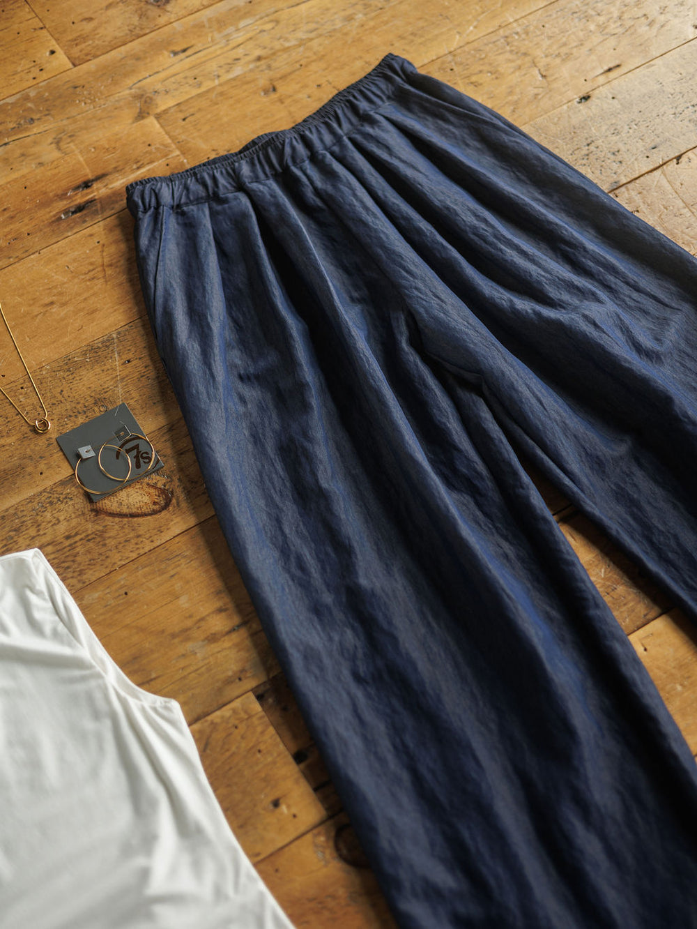 The Meridan high waisted wide leg pants by Savannah Sevens western life{&}style in dark denim blue with an elastic waistband are laid out on a wooden floor beside a white shirt, gold necklace, and gold hoop earrings.