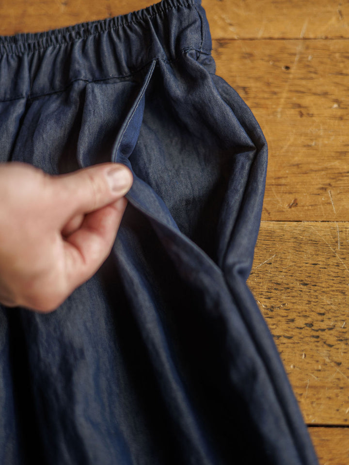A hand opens the side pocket of The Meridan, high waisted wide leg dark denim blue pants from Savannah Sevens western life{&}style, made from a cotton blend and lying on a wooden surface.