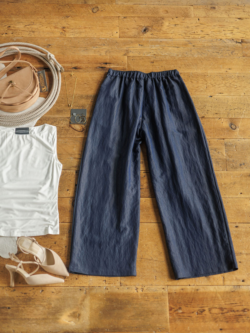 A flat lay of The Meridan by Savannah Sevens western life{&}style: dark denim blue high-waisted wide-leg cotton blend pants, a white sleeveless top, beige heeled sandals, tan round bag, gold necklace, and round sunglasses on a wooden floor.