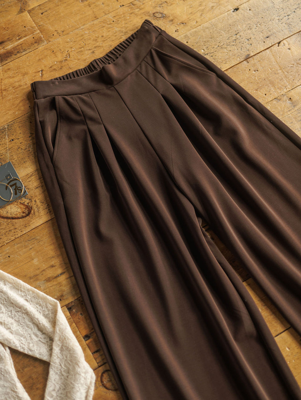 The Kline wide leg pants by Savannah Sevens western life{&}style, featuring dark chocolate pleats and an elastic waistband, are displayed flat on a rustic wooden floor beside a cream lace garment and a clothing tag.