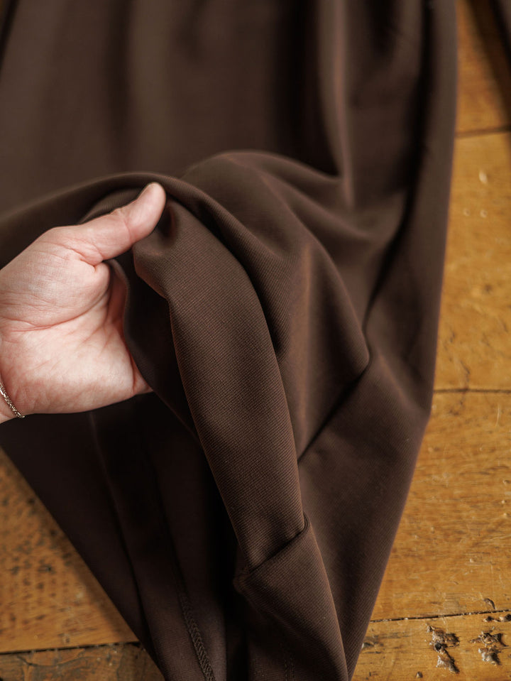 A hand gently lifts a section of The Kline soft, dark chocolate wide leg pants from Savannah Sevens western life{&}style, crafted from tri-blend fabric and draped over a wooden surface.