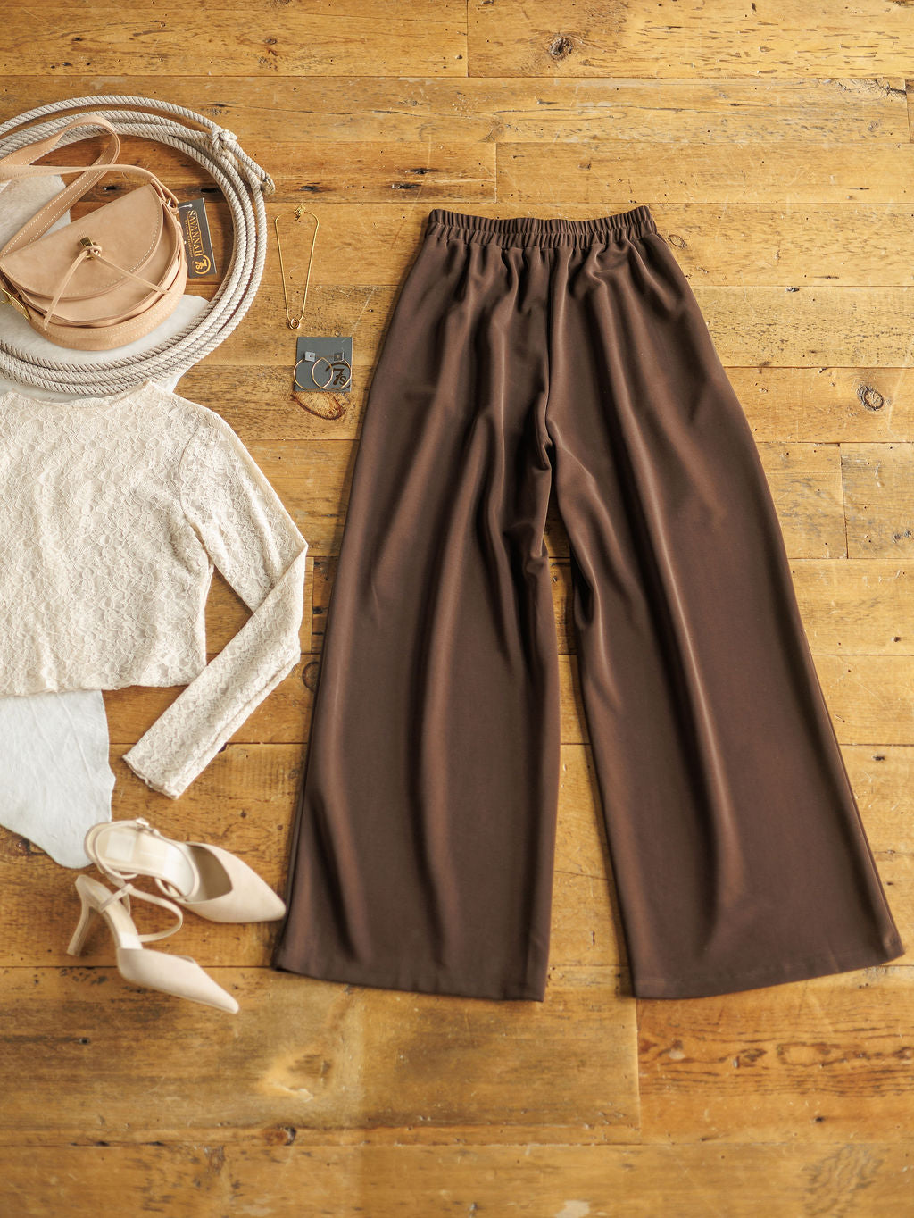 Flat lay of The Kline beige sweater by Savannah Sevens western life{&}style, paired with dark chocolate wide leg pants, beige high heels, a round beige bag, a long necklace, and sunglasses on a wooden floor.