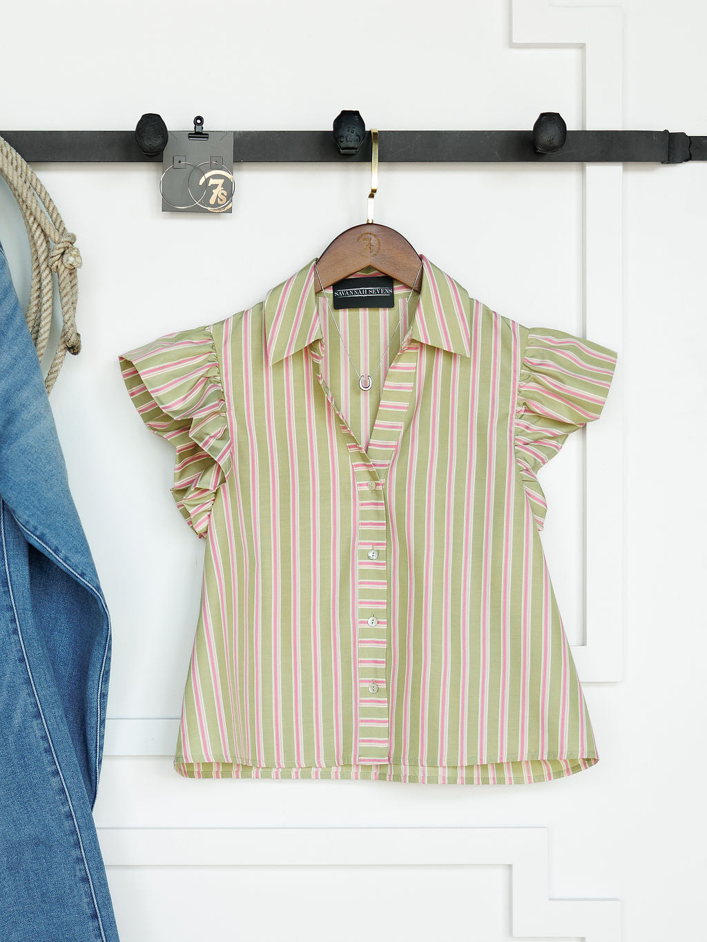 The Rayas by Savannah Sevens western life{&}style—a button-up top with pink and green vertical stripes and flutter sleeves—hangs on a wooden hanger against a white wall, next to a blue denim garment.