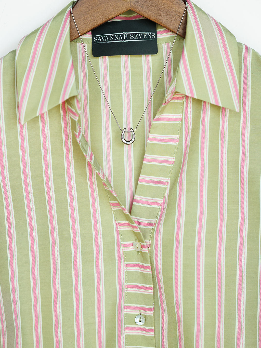 A close-up of The Rayas blouse by Savannah Sevens western life{&}style shows pink and white vertical stripes, buttoned up on a hanger, paired with a silver horseshoe pendant necklace.