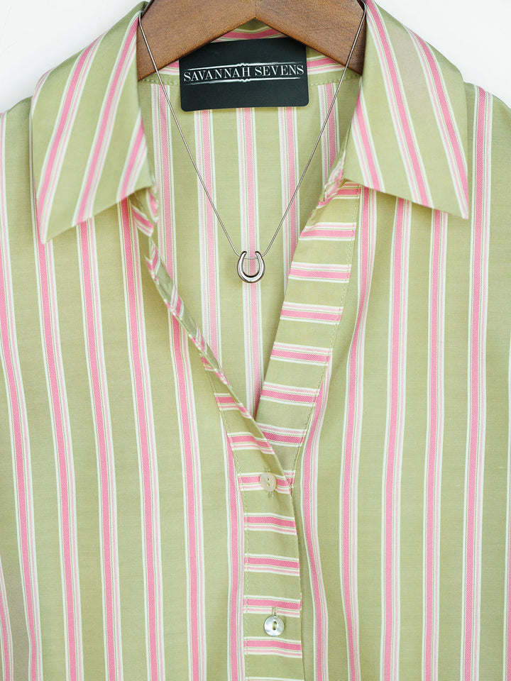 A close-up of The Rayas blouse by Savannah Sevens western life{&}style shows pink and white vertical stripes, buttoned up on a hanger, paired with a silver horseshoe pendant necklace.