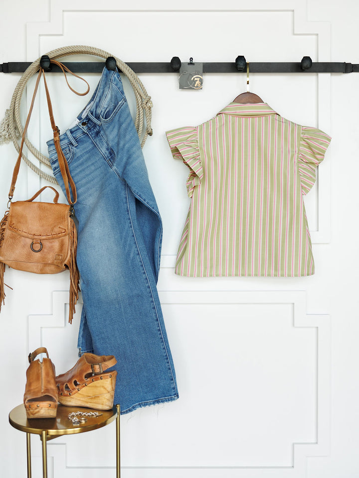 The Rayas striped button-up top by Savannah Sevens western life{&}style is styled with blue jeans, a tan leather bag, and wooden platform sandals, all displayed on a white wall with molding. A rope and small lock hang on the black rack.