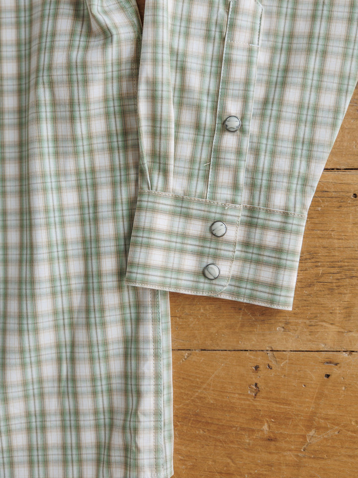 The Eastwood men's plaid snap shirt by Savannah Sevens western life&style, in green and white, lies on a wooden surface. Its features include a double-button cuff, sleeve placket with extra button, and moisture-wicking comfort fabric.