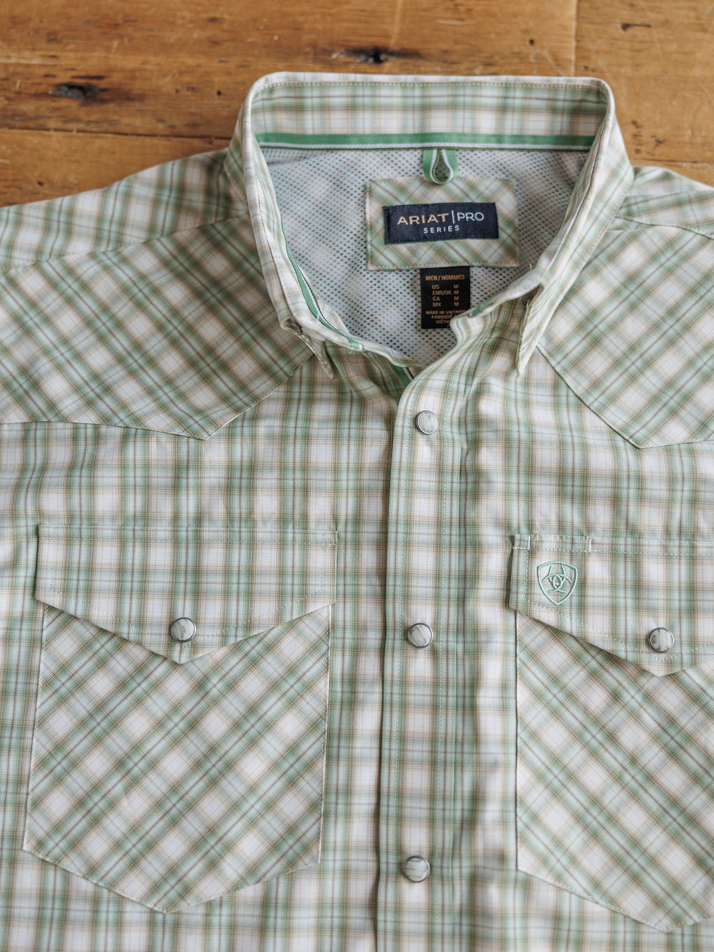 The Eastwood men's shirt by Savannah Sevens western life{&}style features a light green, cream, and tan plaid, two front pockets, silver snaps, logo on the left pocket, moisture-wicking fabric, displayed flat on a wooden surface.