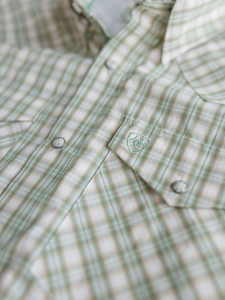 A close-up of Savannah Sevens western life{&}style's The Eastwood {Men's} plaid snap shirt in light green and beige, featuring pearl snaps, a front pocket logo, and moisture-wicking fabric for comfort.