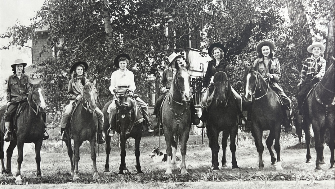 Six people in cowboy hats sit on horses in a row outdoors, framed by trees and a building. The group smiles and poses for the black-and-white photo, capturing a classic western moment.