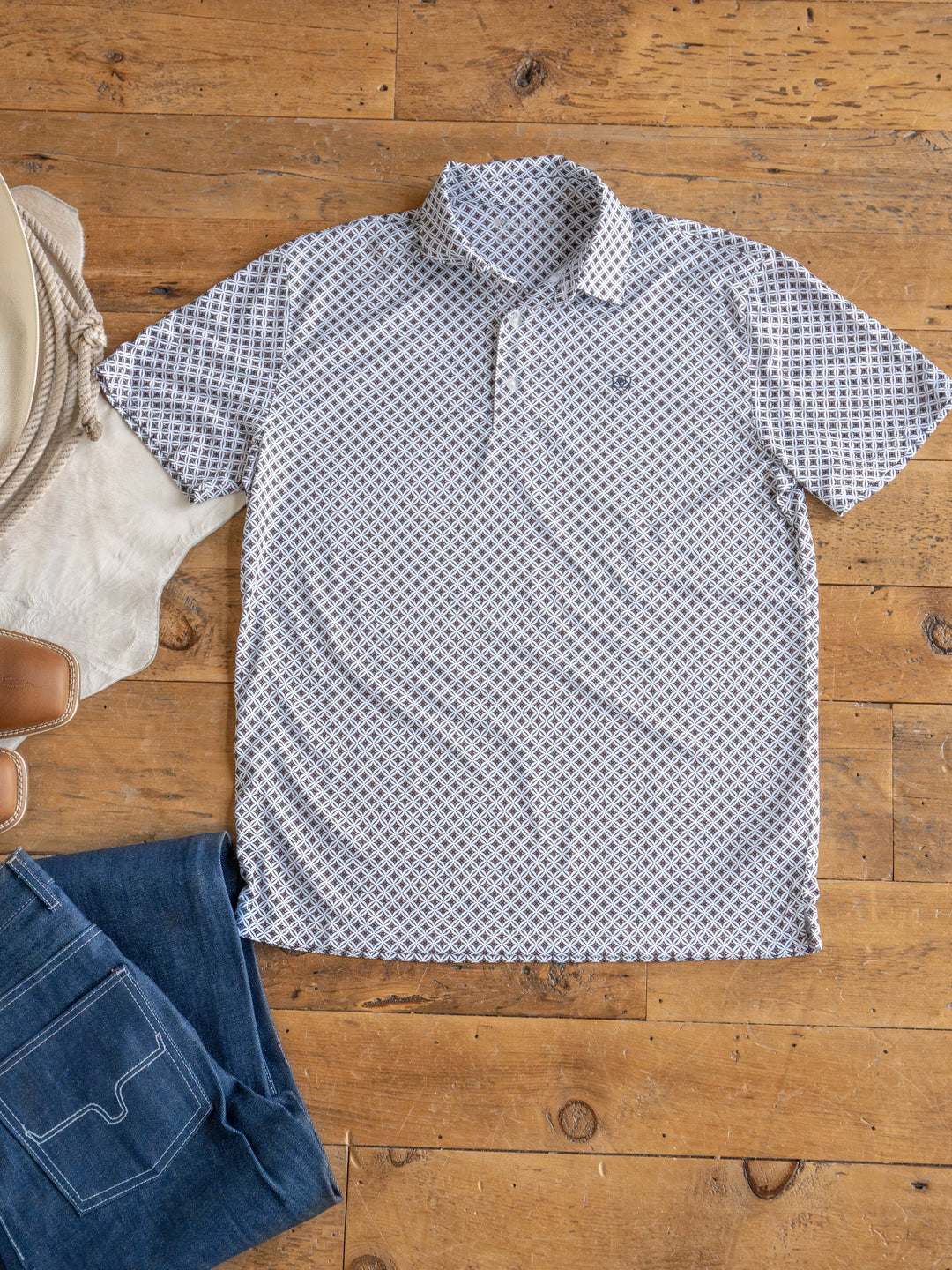 The Lyman men's polo by Savannah Sevens western life{&}style, featuring moisture-wicking fabric and a pattern, is laid flat on a wooden floor next to folded blue jeans, brown boots, a coiled rope, and a white fur rug.