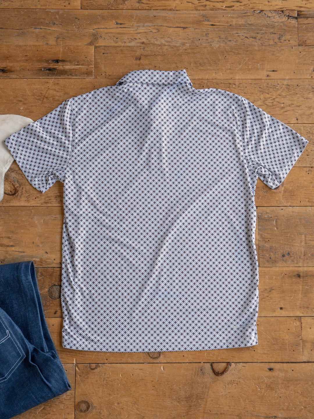 The Lyman Men's polo by Savannah Sevens western life&style, featuring a blue and red geometric print and moisture-wicking fabric, is laid flat with blue jeans on a wooden floor and a touch of white fabric peeking in the corners.