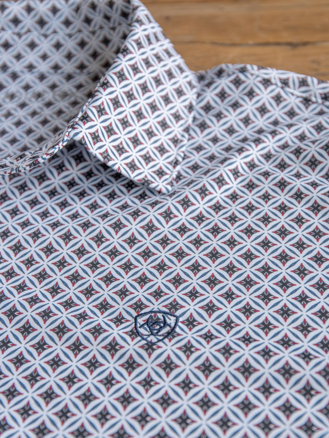 A close-up of The Lyman men's quarter button polo by Savannah Sevens western life&style, with a pointed collar and a geometric design in white, black, and light blue, plus a small embroidered logo on the chest, lying on wood.