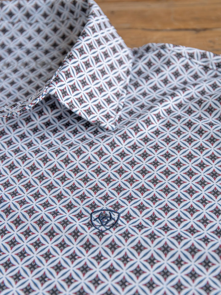 A close-up of The Lyman men's quarter button polo by Savannah Sevens western life&style, with a pointed collar and a geometric design in white, black, and light blue, plus a small embroidered logo on the chest, lying on wood.