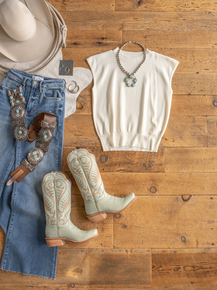 The Hesston by Savannah Sevens western life{&}style: a Western-inspired young contemporary cream cap sleeve top, blue jeans with a decorative belt, light cowboy boots, cowboy hat, hoop earrings, and a turquoise necklace displayed on a wooden floor.