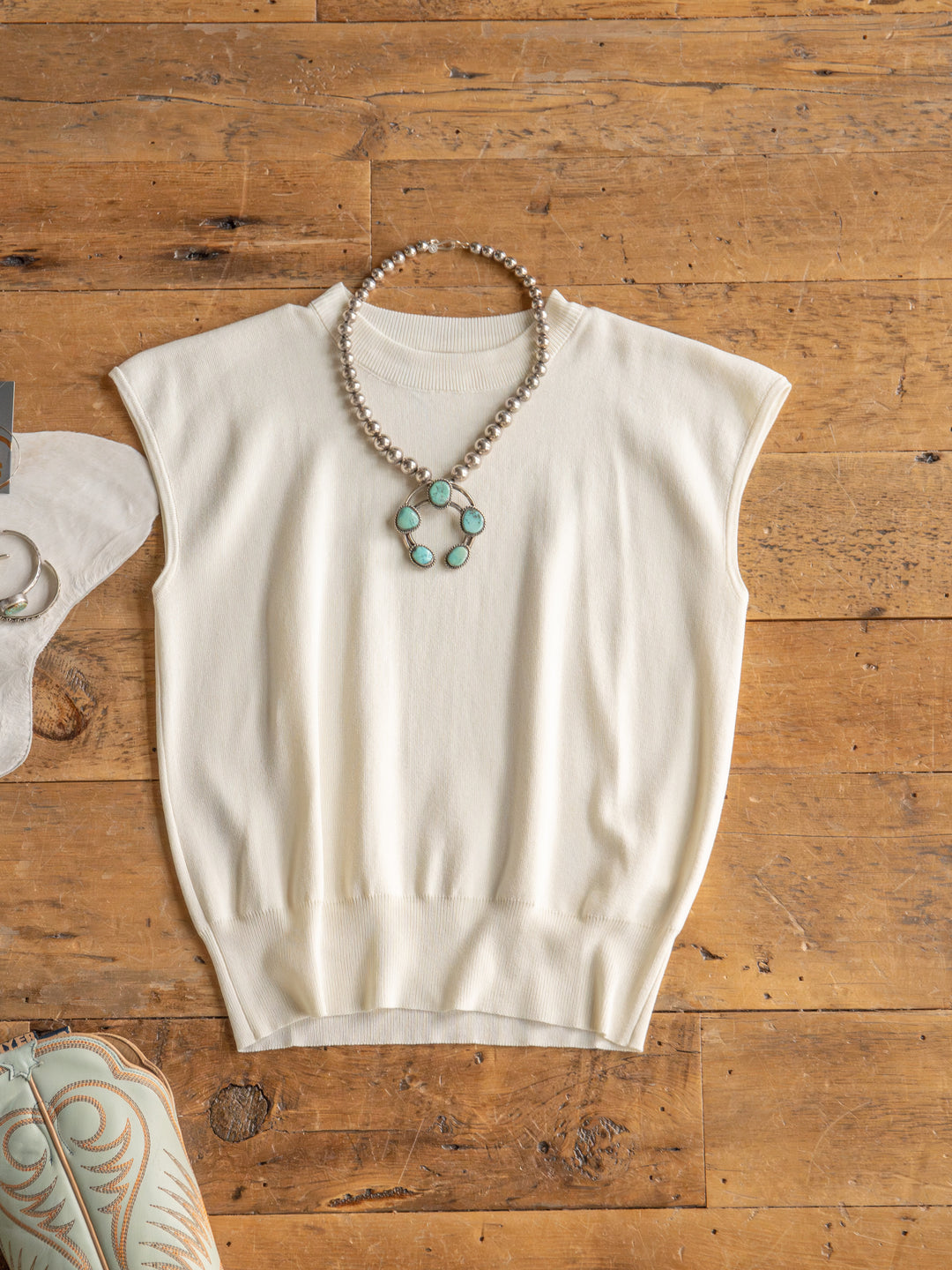 The Hesston by Savannah Sevens western life{&}style, a cream cap sleeve woven top, is laid flat with a pearl necklace and turquoise pendant.