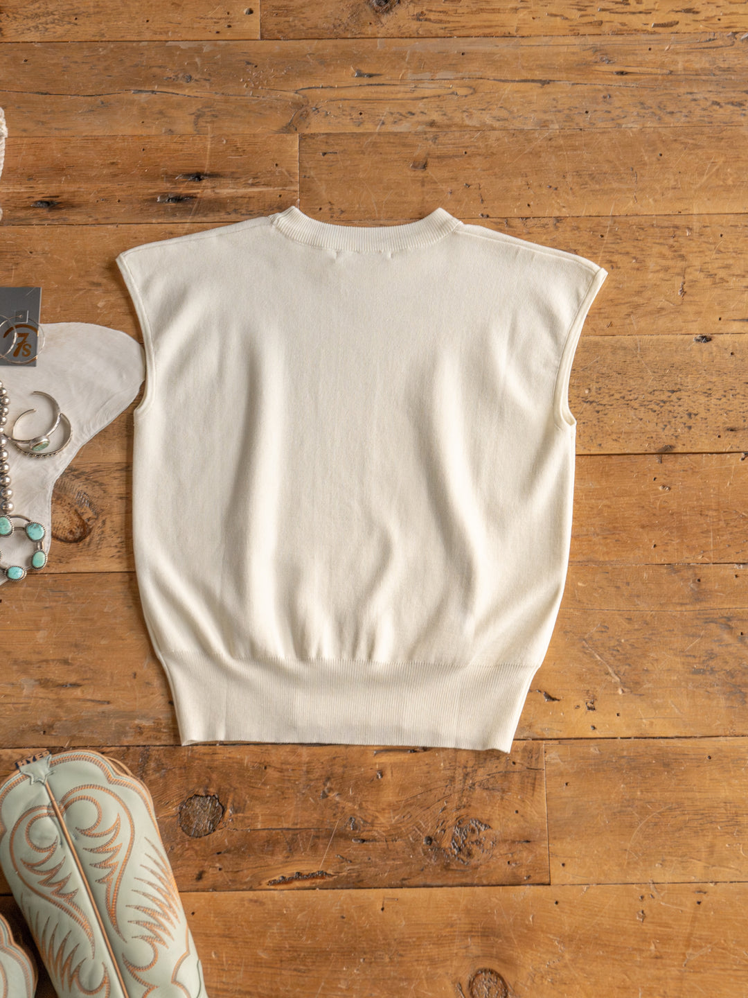 The Hesston by Savannah Sevens western life{&}style is a sleeveless cream cap sleeve top in soft woven fabric, shown flat on rustic wood with silver jewelry and a patterned accent. Features young contemporary sizing for a modern vibe.