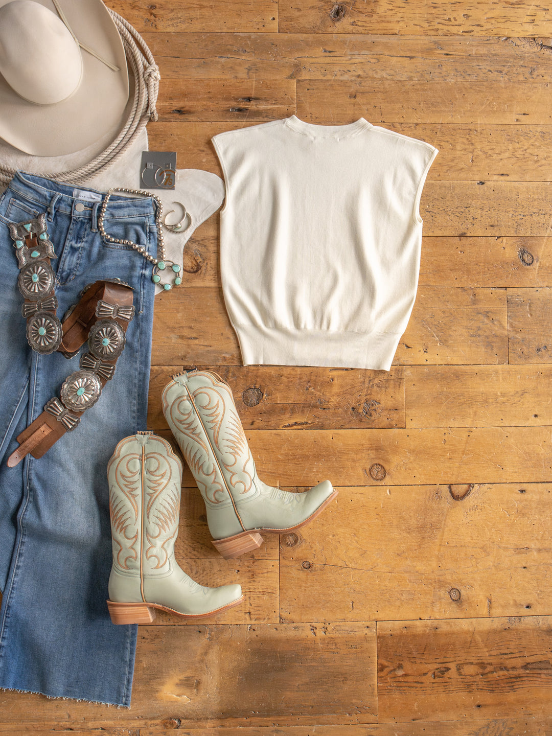 The Hesston by Savannah Sevens western life{&}style: cream cap sleeve top, cowboy hat, turquoise boots, blue denim skirt, tooled leather and concho belts, beaded bracelets, earrings—Western-inspired flat lay in young contemporary sizing.