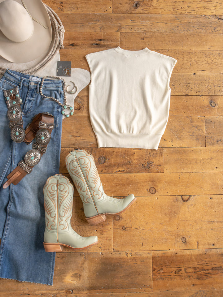 The Hesston by Savannah Sevens western life{&}style: cream cap sleeve top, cowboy hat, turquoise boots, blue denim skirt, tooled leather and concho belts, beaded bracelets, earrings—Western-inspired flat lay in young contemporary sizing.