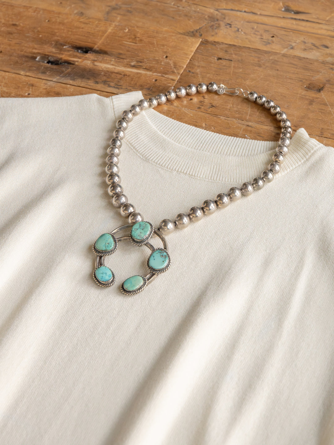 The Hesston by Savannah Sevens western life{&}style, a cream cap sleeve top in soft woven fabric, is displayed on wood with a silver beaded necklace featuring five turquoise stones.