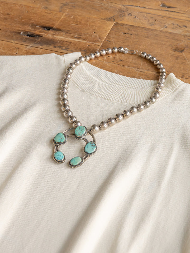 The Hesston by Savannah Sevens western life{&}style, a cream cap sleeve top in soft woven fabric, is displayed on wood with a silver beaded necklace featuring five turquoise stones.
