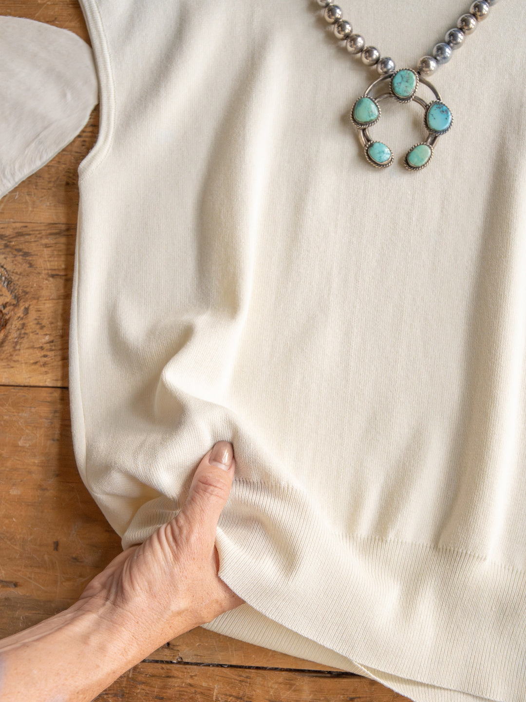 A hand gently pinches the hem of The Hesston cream cap sleeve top by Savannah Sevens western life{&}style, crafted from soft woven fabric and shown on wood with a turquoise and silver necklace placed on it.