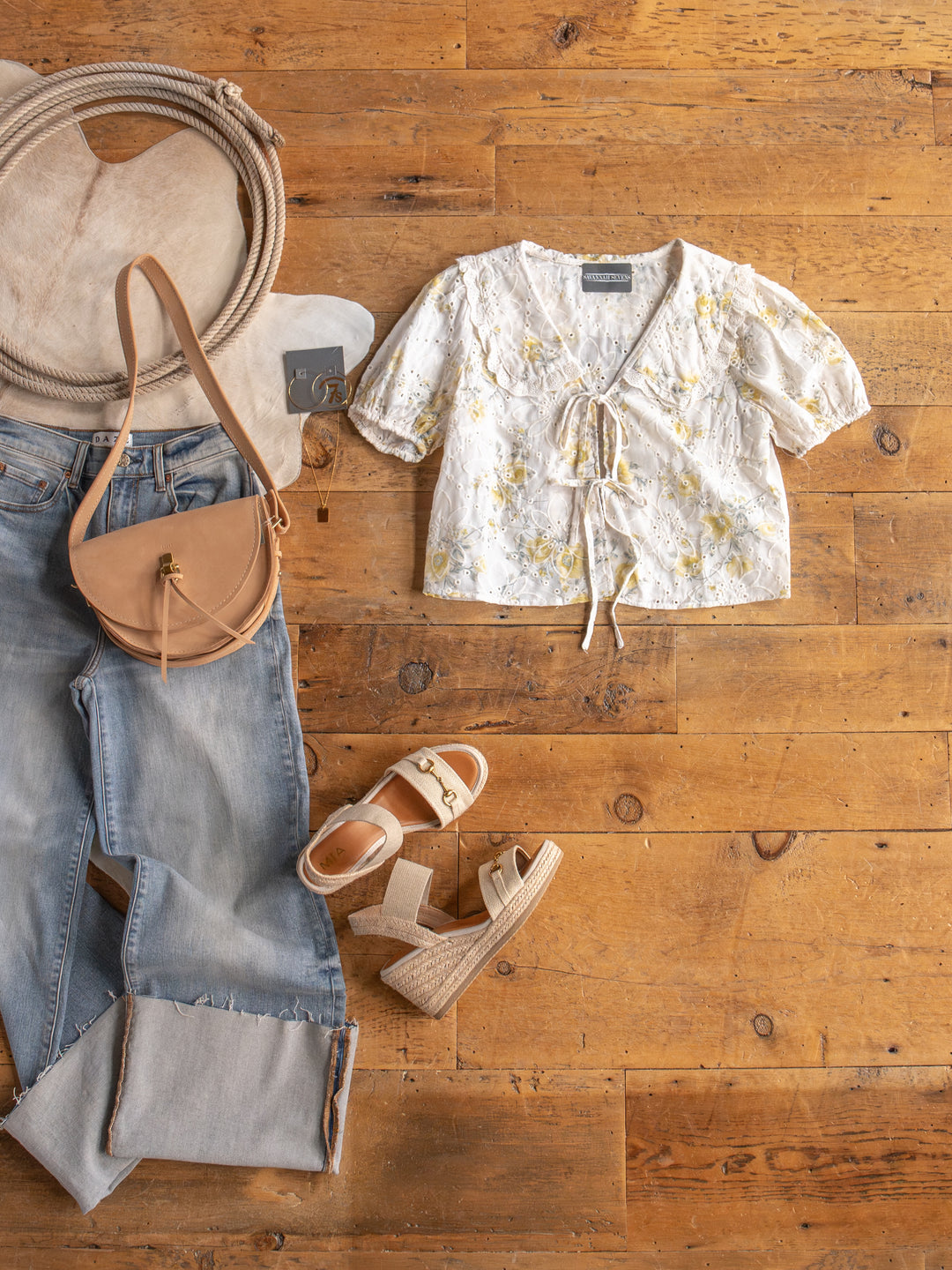 Flat lay of The Silsbee floral front tie top by Savannah Sevens western life{&}style, light wash jeans, beige wedge sandals, tan crossbody bag, and round hat with rope accent on a wooden floor.