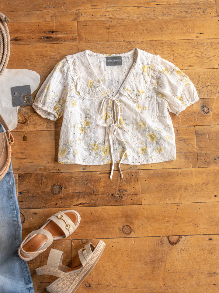 The Silsbee by Savannah Sevens western life{&}style, a sheer embroidered top with yellow floral details, is displayed on a wooden floor beside beige sandals, blue jeans, a tan bag, earrings, and a necklace.