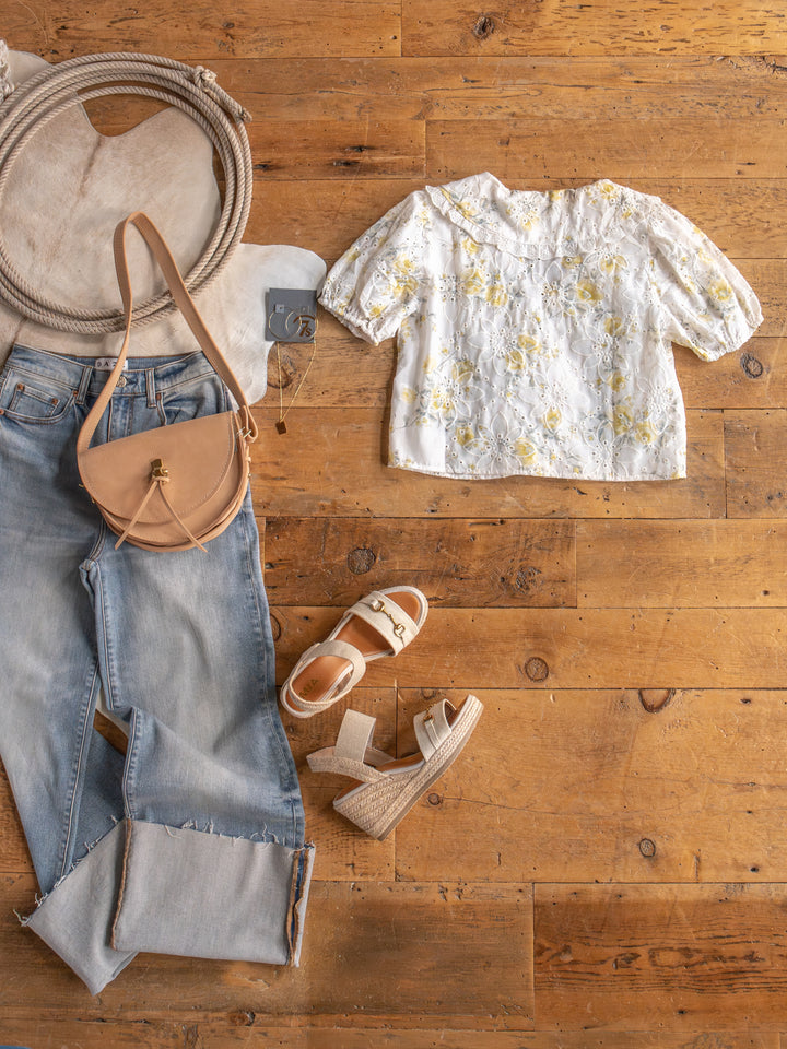Flat lay of a casual women’s outfit from Savannah Sevens western life{&}style featuring The Silsbee: light blue jeans, floral front tie top, tan crossbody bag, cream heeled sandals, and tan flat sandals.