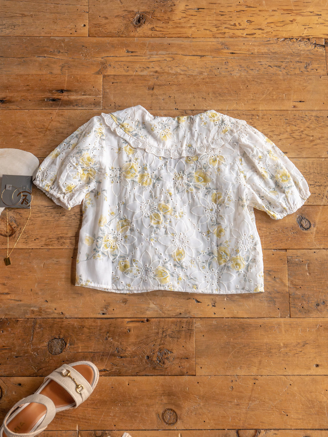 The Silsbee white floral front tie top with yellow patterns and puffed sleeves by Savannah Sevens western life{&}style is laid flat on a wooden floor, next to beige sandals and a gold necklace.