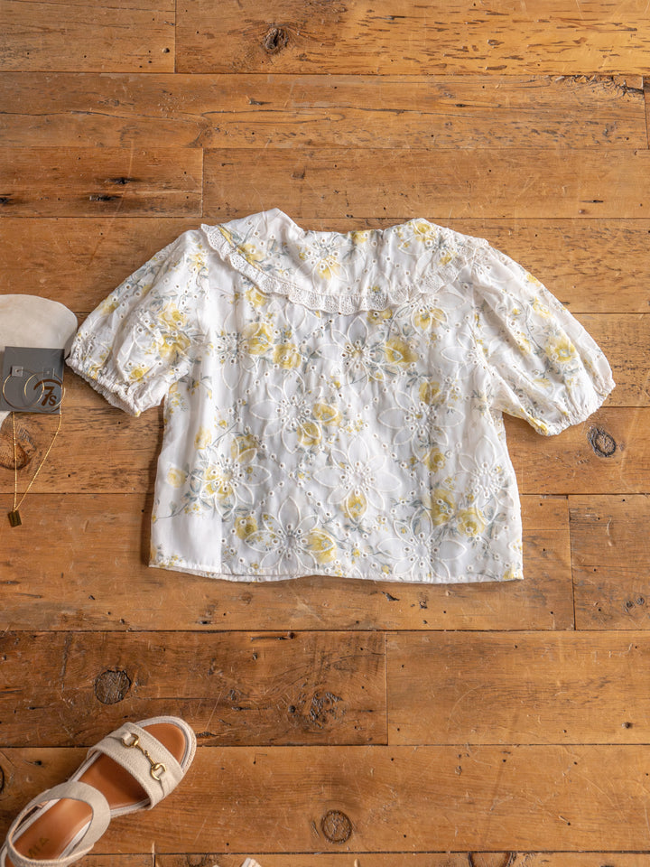 The Silsbee white floral front tie top with yellow patterns and puffed sleeves by Savannah Sevens western life{&}style is laid flat on a wooden floor, next to beige sandals and a gold necklace.