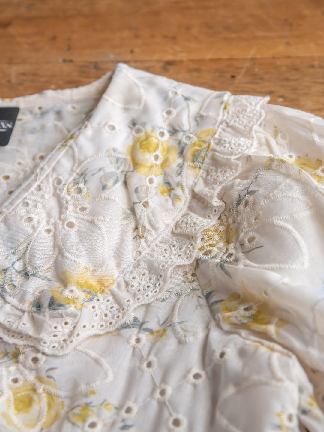 The Silsbee by Savannah Sevens western life{&}style: a sheer embroidered top with yellow floral accents and lace on the collar and shoulders, displayed on a wooden surface.