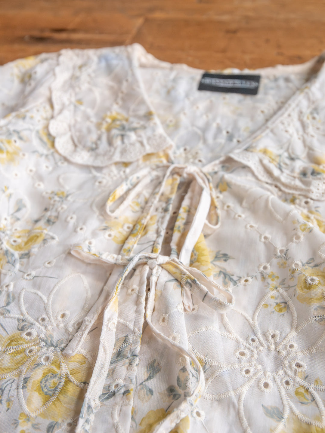 A close-up of The Silsbee by Savannah Sevens western life{&}style—a sheer embroidered floral top with a scalloped lace collar and front tie neckline, displayed on a wooden surface.