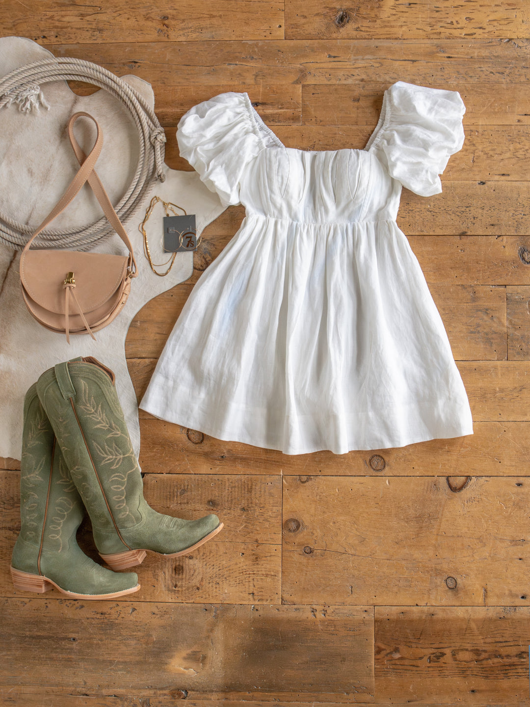 The Southampton white linen puff sleeve dress by Savannah Sevens western life{&}style is styled with sage green cowboy boots, a tan crossbody bag, sunglasses, a gray-cased phone, and a cowhide rug on a wooden floor.