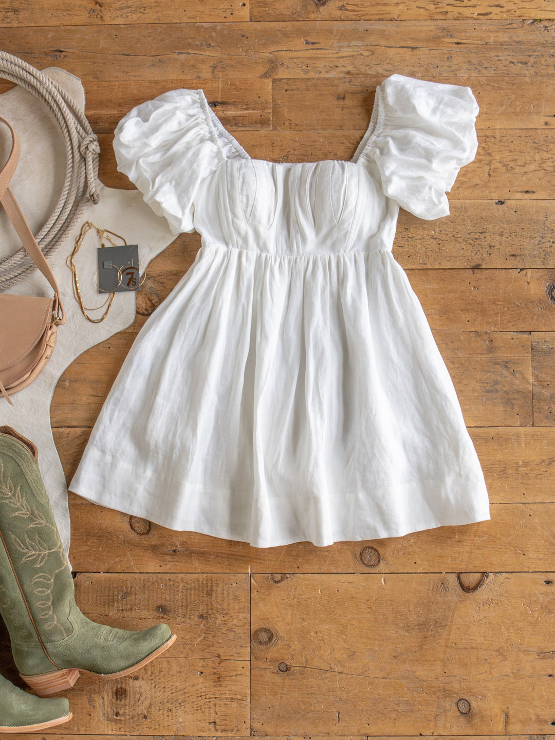 The Southampton white linen puff sleeve dress by Savannah Sevens western life{&}style is laid flat on a wooden floor, styled with green embroidered cowboy boots, a tan crossbody bag, a rope coil, and a gold tag necklace.