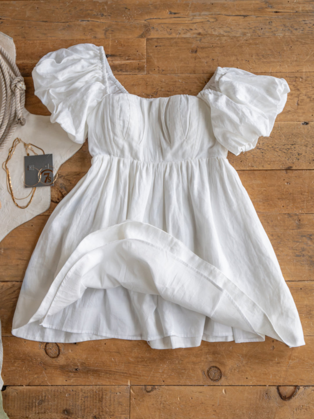 The Southampton by Savannah Sevens western life{&}style is a white linen puff sleeve mini dress with a fitted bodice, laid on a wooden floor with accessories and tags, showcasing its lightweight, summery charm.