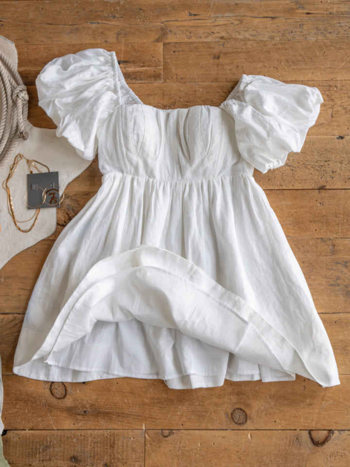 The Southampton by Savannah Sevens western life{&}style is a white linen puff sleeve mini dress with a fitted bodice, laid on a wooden floor with accessories and tags, showcasing its lightweight, summery charm.