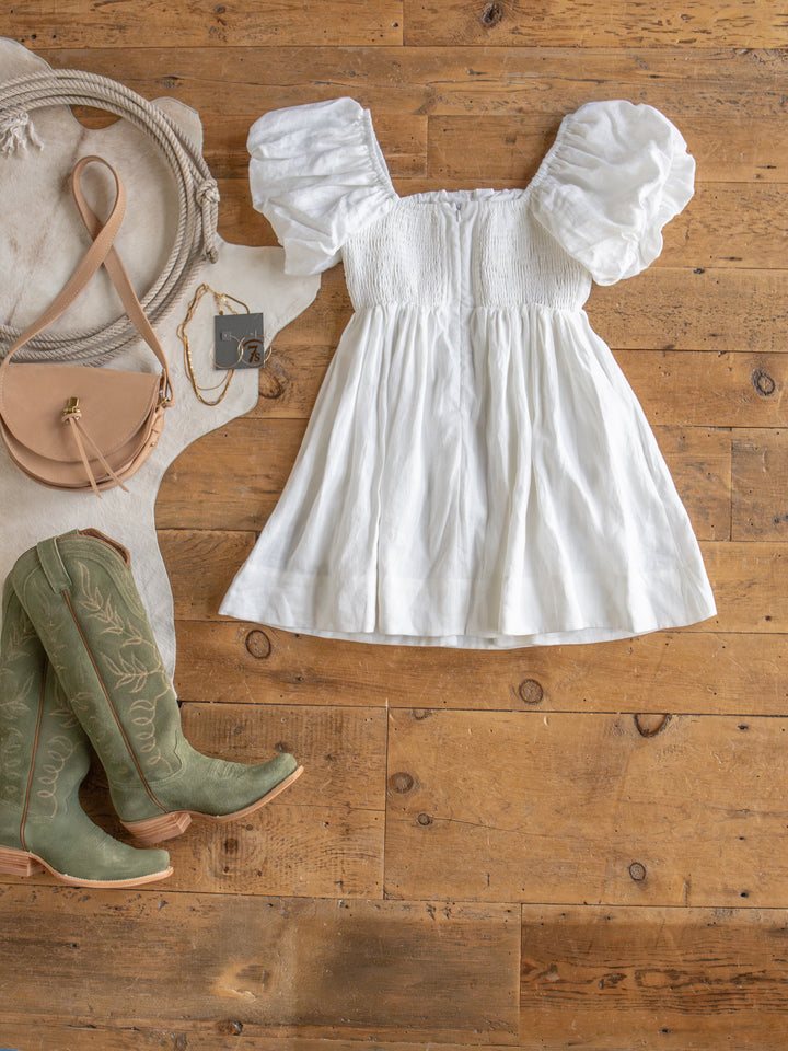 The Southampton linen puff sleeve dress by Savannah Sevens western life{&}style is laid out on a wooden floor, next to a tan purse and green embroidered cowboy boots.