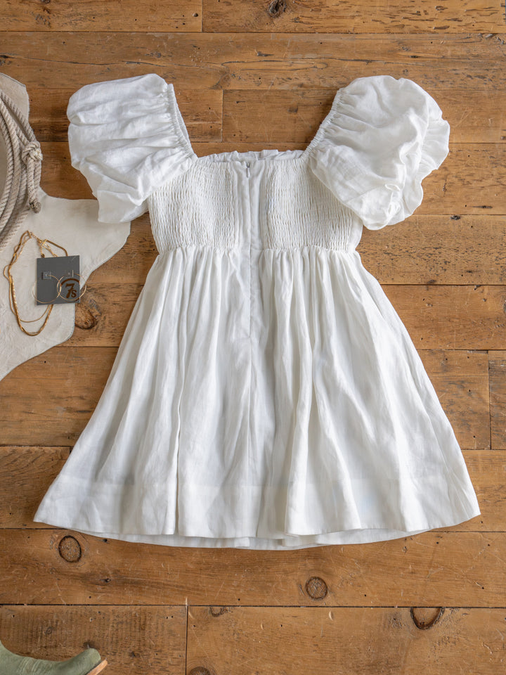 The Southampton white linen puff sleeve dress by Savannah Sevens western life{&}style is laid flat on a wooden floor, surrounded by a partial green shoe, rope bag, and gold necklace.