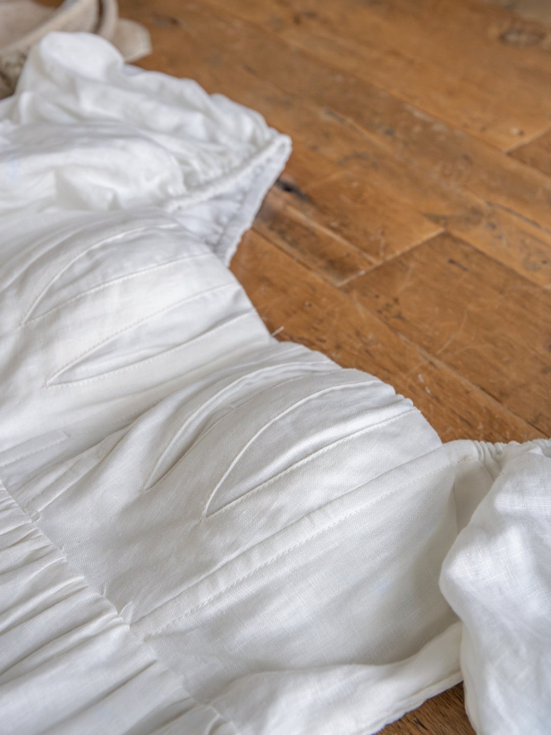 A close-up of The Southampton by Savannah Sevens western life{&}style—a white linen puff sleeve dress with ruched details, light textured fabric, and gentle folds—lying on a wooden floor.