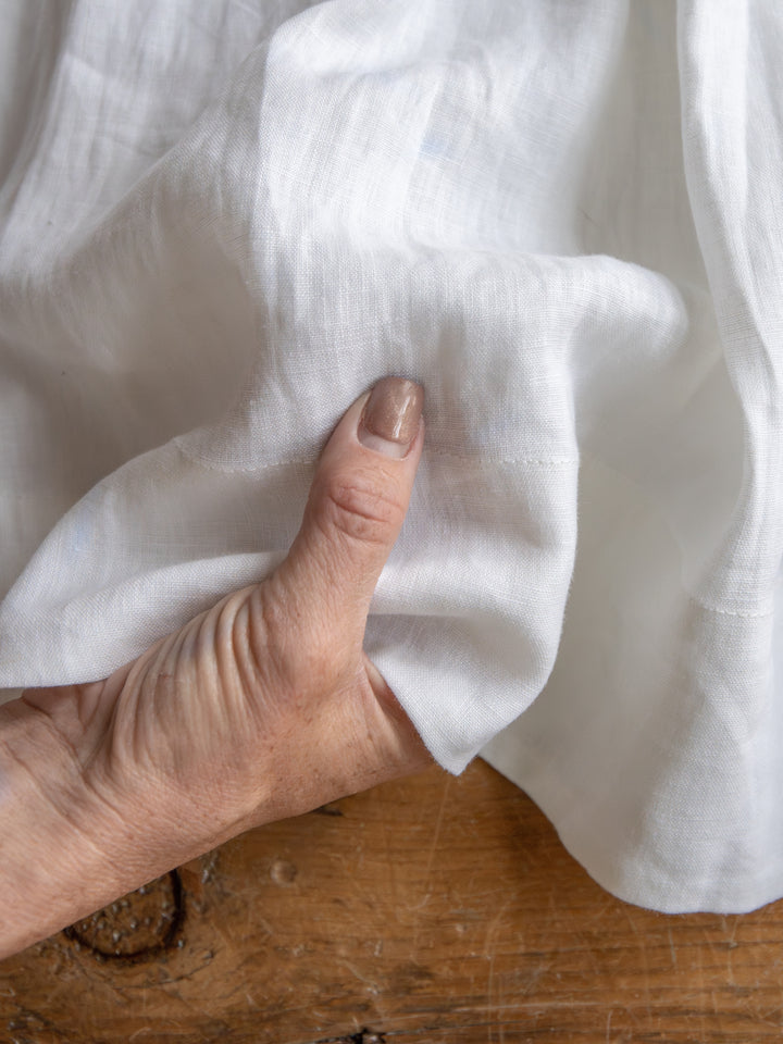 A hand gently holds The Southampton white linen puff sleeve dress by Savannah Sevens western life{&}style against a wooden surface.