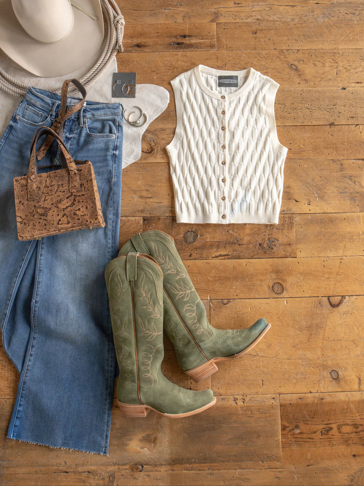 Flat lay of The Patero, a textured sleeveless knit top by Savannah Sevens western life{&}style, paired with jeans, sage green cowboy boots, handbag, beige wide-brim hat, and silver hoop earrings on a wooden floor.
