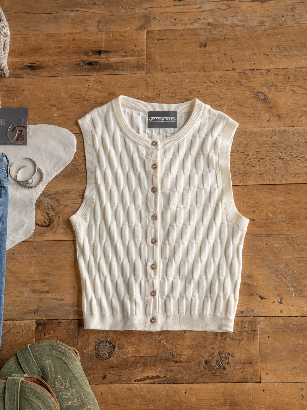 The Patero by Savannah Sevens western life{&}style is a cream, sleeveless textured knit top with a button-up design, shown flat on wood, with parts of a white hat, green shoes, and blue jeans nearby.