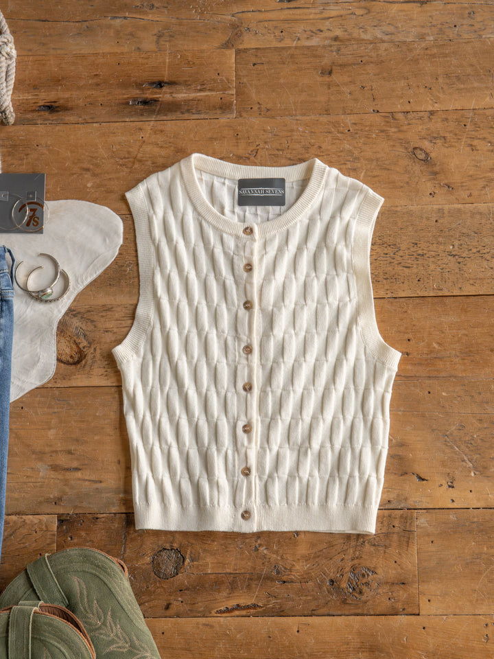 The Patero by Savannah Sevens western life{&}style is a cream, sleeveless textured knit top with a button-up design, shown flat on wood, with parts of a white hat, green shoes, and blue jeans nearby.