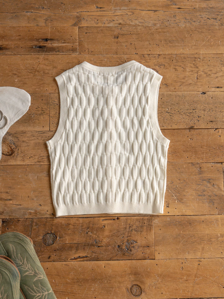 The Patero by Savannah Sevens western life{&}style is a cream-colored sleeveless knit vest with an oval textured pattern, displayed flat on a rustic wooden floor. Partial views of a green shoe and white accessory are visible at the edges.