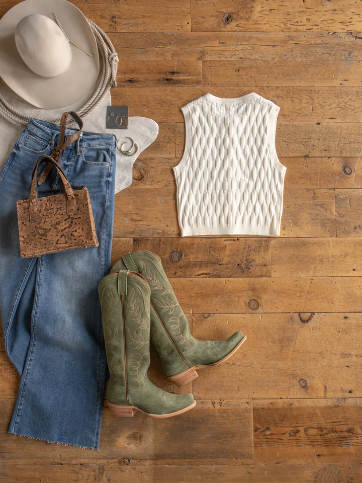 Flat lay of The Patero boots by Savannah Sevens western life{&}style, styled with a light beige hat, textured sleeveless top, light blue button-up jeans, cork handbag, and silver hoop earrings on a wooden floor.